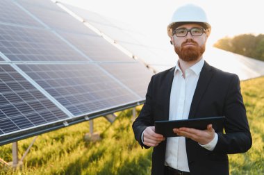 Sert bir şapka takan ve tablet kullanan yatırımcı, canlı bir günbatımında güneş paneli çiftliğini inceleyerek yenilenebilir enerji yeniliklerini vurguluyor.