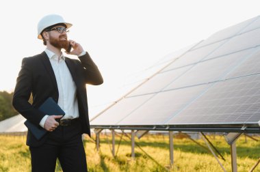 Gün batımında güneş panelleri olan sürdürülebilir bir enerji santralini incelerken tablet tutan ve akıllı telefon kullanan mühendis