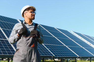 Elektrik mühendisi, açık mavi gökyüzünün altındaki büyük bir fotovoltaik fabrikayı incelerken iş kıyafetlerini ayarlıyor.
