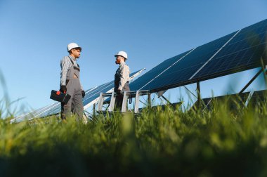 İki mühendis, güneşli bir günde yeşil bir alana fotovoltaik paneller kurarak bir güneş enerjisi santrali üzerinde çalışıyorlar.