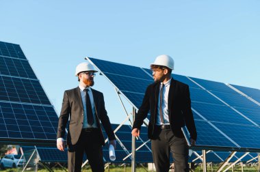 Giysi ve şapka giyen iki mühendis güneş enerjisi istasyonunda güneş panelinin yanında yenilenebilir enerji geliştirmeyi tartışıyorlar.