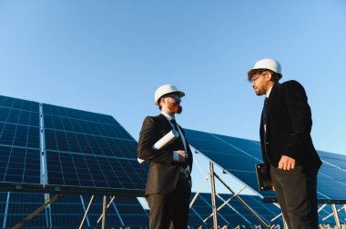 İki mühendis, ellerinde planlar olan ve güneş enerjisi istasyonunda güneş panellerinin yanında tartışan zırhlar ve sert şapkalar takıyor.