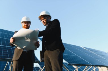 Şapkalı iki mühendis bir güneş paneli çiftliğinde yenilenebilir enerji projesi üzerinde çalışıyorlar.