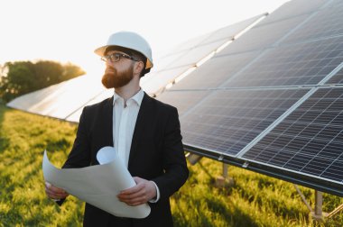 Kask takan ve elinde bir plan tutan mühendis güneş paneli çiftliğini gün batımında inceleyerek yenilenebilir enerji yeniliği ve sürdürülebilirliği vurguluyor.