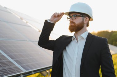 Takım elbiseli bir yatırımcı güneş paneli çiftliğini gün batımında teftiş ederken kaska dokunuyor.