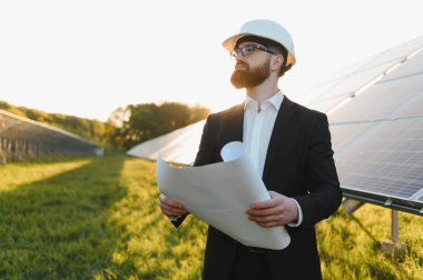 Güneş enerjisi santralini gün batımında incelerken takım elbiseli bir yatırımcı elinde planlar tutuyor.