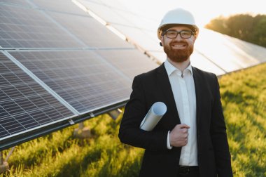 Güneş enerjisi santralinin planlarını tutarken gülümseyen mühendis, çarpıcı bir gün batımının sıcaklığında güneşleniyor, yenilik ve sürdürülebilirliği somutlaştırıyor.