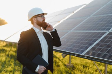 Yatırımcı elinde bir tablet tutarken ve akıllı telefon kullanırken açık havada gün batımında güneş panellerini inceliyor.