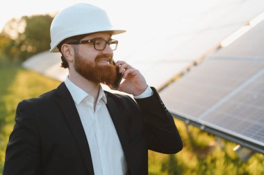 Yatırımcı gün batımında güneş panellerinin yanında akıllı telefon kullanırken takım elbise giyip gülümsüyor.