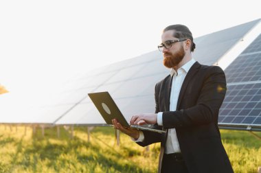 Laptop kullanan iş adamı, yeşil alandaki güneş panellerini denetliyor, yenilenebilir enerji projesi üzerinde çalışıyor.
