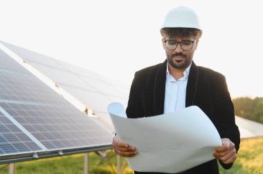 Hintli mühendis güneş enerjisi santralinde dururken baret takıp planı analiz ediyor.