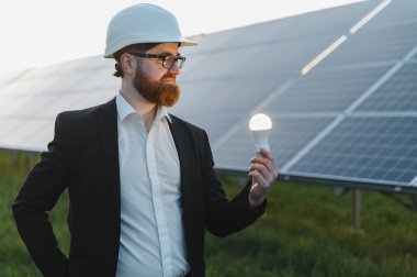 Yenilenebilir enerji ve yeşil yatırımları sembolize eden, güneş enerjisi santralinin önünde bir ampul tutan mühendis.