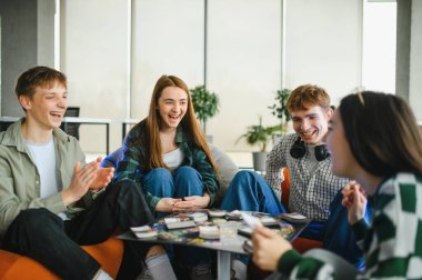 Dört neşeli genç birlikte vakit geçiriyorlar, masa oyunu oynuyorlar ve gülüyorlar, rahat puf koltuklarda oturuyorlar.