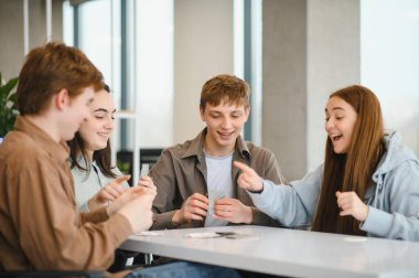 Dört neşeli öğrenci bir masanın etrafında oturuyor, kağıt oynuyor ve okulda birlikte vakit geçiriyorlar.