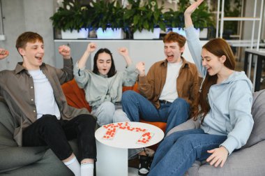 Dört neşeli genç, küçük bir masanın etrafında armut koltukta oturmuş masa oyunu oynuyor ve kollarını kaldırarak heyecanlarını dile getiriyorlar.