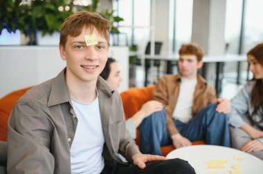 Alnında yapışkan bir notla mutlu genç adam bil bakalım modern ofis ortamında arkadaşlarıyla kim oynuyor?