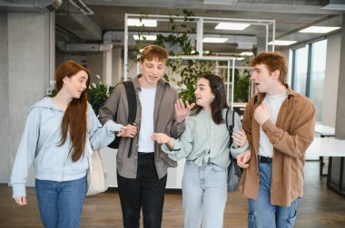 Sırt çantalı dört mutlu üniversite öğrencisi üniversite koridorunda yürüyüp sohbet ediyorlar.