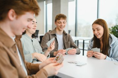 Dört mutlu kolej öğrencisi, beyaz bir masanın etrafında oturmuş, bir üniversitenin dinlenme odasında kağıt oynuyor ve birlikte eğleniyorlar.