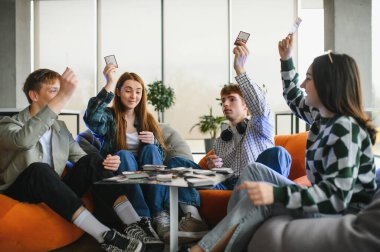 Dört neşeli genç yetişkin, boş zamanlarının tadını çıkarıyorlar, modern bir dairede puf koltuklarda oturmuş kart oynuyorlar.