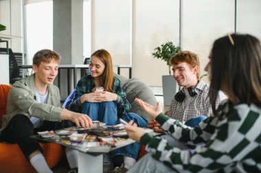 Dört üniversite öğrencisi gülüyor ve boş zamanlarının keyfini çıkarırken canlı bir üniversite salonunda kart oynuyor, arkadaşlığı ve eğlenceyi kucaklıyor.