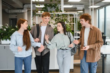 Dört mutlu öğrenci bir üniversite koridorunda birlikte yürüyüp konuşuyorlar, sırt çantaları taşıyorlar ve arkadaşça sohbet ediyorlar.