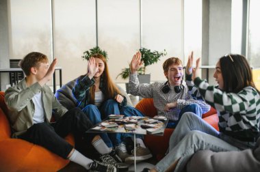 Dört neşeli genç yetişkin puf minderlerinde oturmuş masa oyunu oynuyor ve bir beşlik çakıyor, sevinçlerini ve heyecanlarını dile getiriyorlar.
