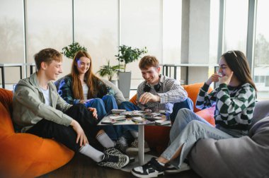 Dört neşeli üniversite öğrencisi armut koltukta oturuyor, kağıt oynuyor ve modern ortak bir alanda eğleniyorlar.