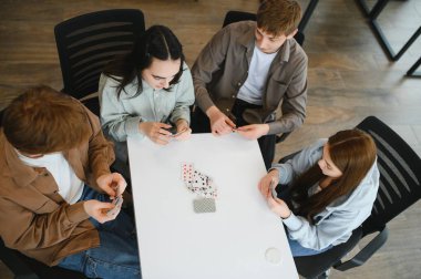 Dört arkadaş beyaz bir masada oturuyor, kağıt oynuyor ve eğleniyorlar, yukarıdan bakıyorlar.