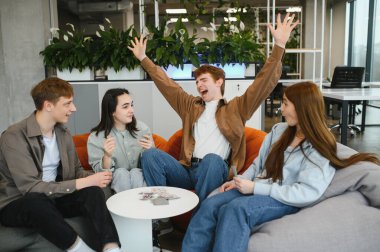 Dört neşeli meslektaşımız kağıt oyunlarının keyfini çıkarırken, bir tanesi modern ofis alanlarında kazanan eli heyecanla kutluyor.