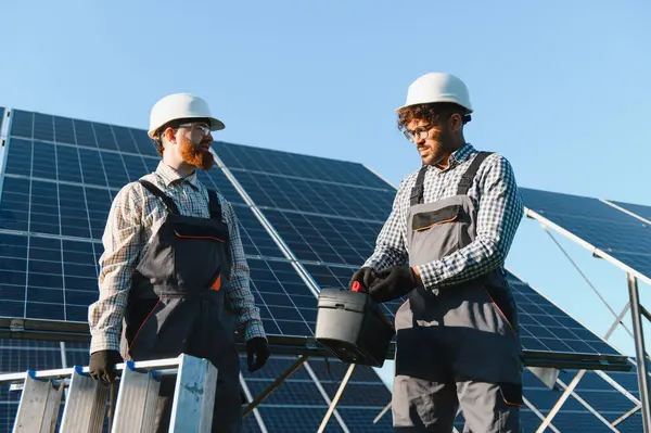 Güneş paneli kurulumunda işbirliği yapan iki mühendis, açık mavi gökyüzünün altındaki bir alet kutusundan aletleri dikkatlice alıyorlar.