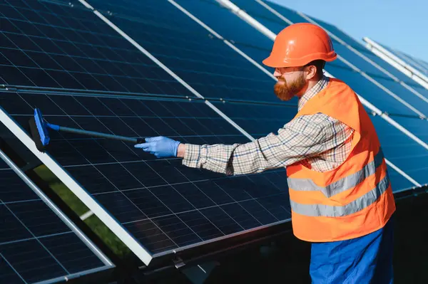 Erkek işçi, güneş panellerini fırçayla temizliyor ve büyük bir güneş enerjisi santralinde maksimum verimi sağlıyor.