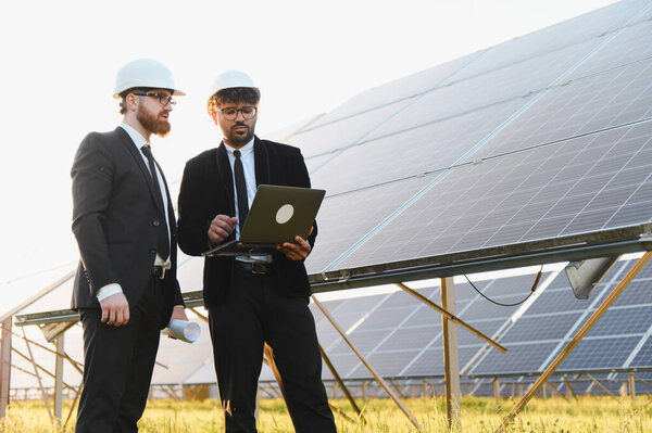 Два инженера с чертежами и ноутбуком, работающими на солнечной электростанции, изучают фотоэлектрические панели и обсуждают возобновляемые источники энергии
