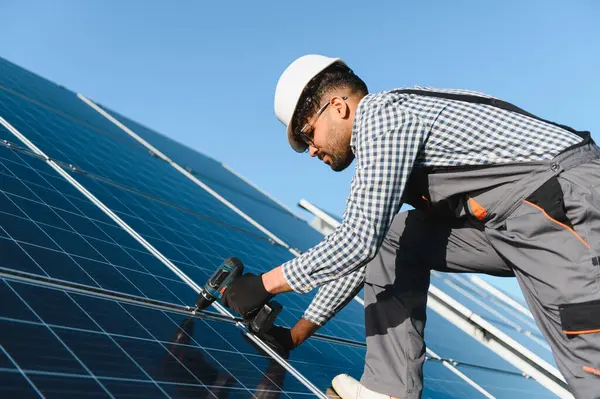 Elektrikçi çatıya fotovoltaik paneller yerleştirmek için tornavida kullanıyor, güvenlik kaskı ve eldiven takıyor, yenilenebilir enerji projesi üzerinde çalışıyor.