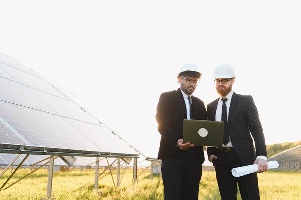 Два инженера в хардхатах и деловых костюмах используют ноутбук и чертежи для анализа солнечных панелей фермы на закате, представляя возобновляемые источники энергии и устойчивости