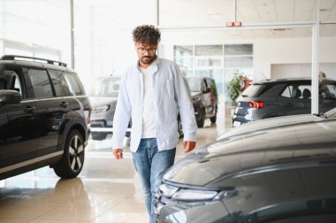 Arap bir adam araba galerisinde yürüyor, seçenekleri değerlendiriyor ve modern sergi salonundan yeni bir araç seçiyor.