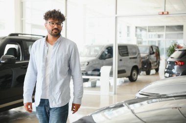 Arap bir adam modern bir araba galerisinde yürüyor, yeni bir araç seçiyor ve çeşitli seçenekleri göz önünde bulunduruyor.