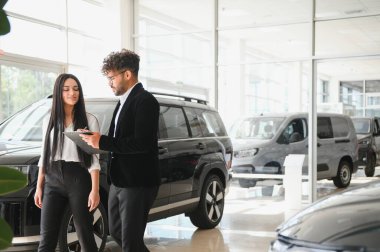 Bir araba galerisinde müşteriye yeni araçlar gösteren satıcı, özellikleri ve finansman seçeneklerini tartışıyor