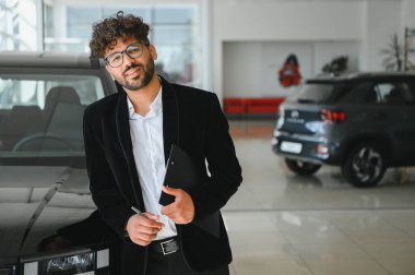 Satıcı elinde bir pano ve kalem tutarken gülümsüyor, galeride bir arabanın yanında duruyor.