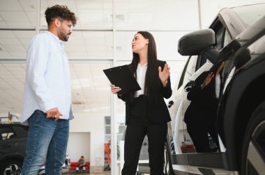 Profesyonel satıcı elinde bir pano var. Otomobil galerisindeki erkek müşteriye yeni bir araba sunuyor.