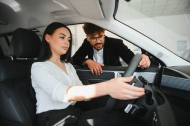 Araba satıcısı, bir galeride sürücü koltuğunda oturan bir kadına yeni bir arabanın özelliklerini anlatıyor.