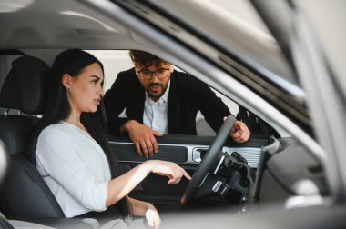 Satıcı, bir araba galerisinde sürücü koltuğunda oturan kadın müşteriye araba özelliklerini açıklıyor.