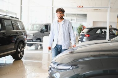 Arap adam yeni bir araba seçiyor, kaputa nazikçe dokunuyor ve parlak, ferah bir araba galerisinde seçenekleri araştırıyor.