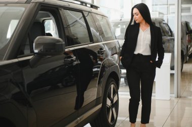 İş kadını, bir araba galerisinde şık siyah bir cipin yanında özgüvenle duruyor, yaklaşan alımını düşünceli bir şekilde düşünüyor.