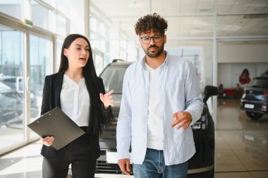 Satıcı kadın elinde bir pano ile modern bir araba galerisindeki müşterisine yeni bir araba sunuyor. Seçenekleri ve finansman detaylarını tartışıyor.