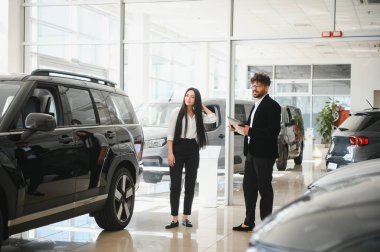 Satıcı, modern bir araba galerisinde bayan bir müşteriye yeni bir SUV sunarken, satın alma sürecini kolaylaştıran bir tablet sunuyor.