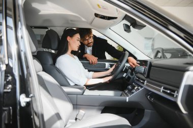 Satıcı, araba özelliklerini bir bayide sürücü koltuğunda oturan bir kadına anlatıyor.