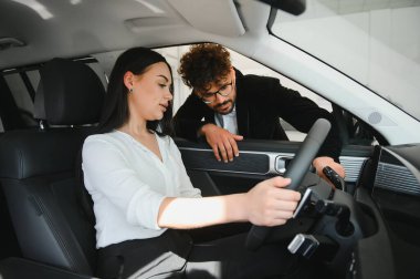 Araba satıcısı, bir araba galerisinde sürücü koltuğunda oturan kadın müşteriye direksiyon fonksiyonlarını açıklıyor.