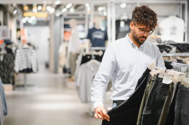 Genç Arap adam bir giyim mağazasında pantolonunu dikkatlice seçiyor, elinde siyah bir elbise tutuyor.