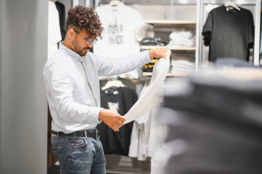 Zarif bir Arap adam şık bir gömleği incelerken modern bir giyim mağazasından alışveriş yapıyor.