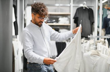 Zarif bir Arap adam modern bir giyim mağazasında alışveriş yaparken şık bir beyaz gömleği inceliyor, modaya uygun bir perakende deneyimin tadını çıkarıyor.